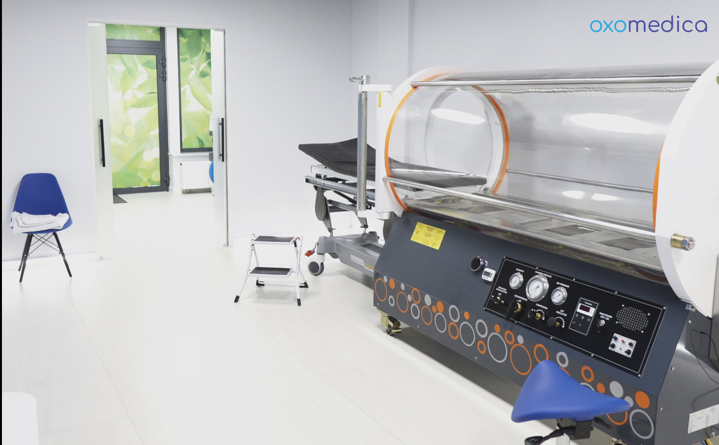Wnętrze kliniki: komora hiperbaryczna Oxomedica z manometrami i leżanką. W tle krzesło i otwarte drzwi z motywem liści. Jasne, sterylne otoczenie.