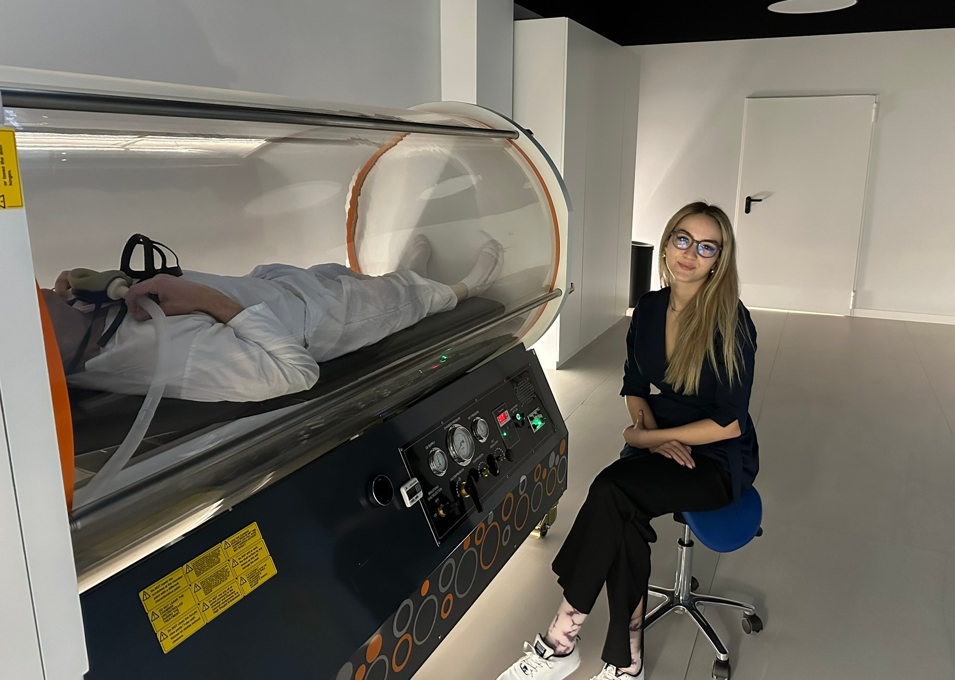 Pacjent w komorze hiperbarycznej z maską tlenową, obok siedzi kobieta w okularach, w tle białe ściany i drzwi. Klinika oferuje zabiegi tlenoterapii hiperbarycznej.