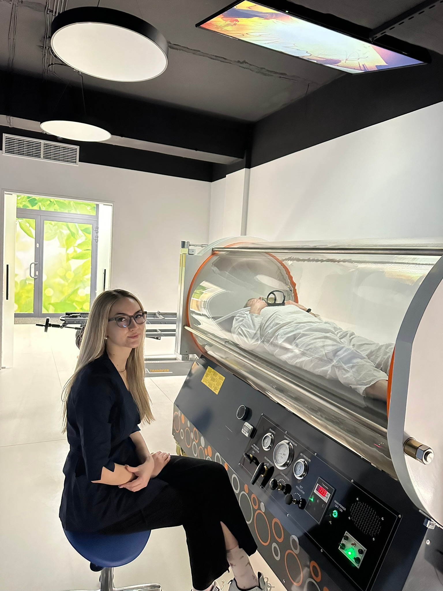 Kobieta siedzi obok komory hiperbarycznej z pacjentem w środku. Widoczne okno, oświetlenie sufitowe i aparatura kontrolna komory.