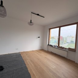 Piotr Kowalczyk P&P Profinish - Układanie drewnianej podłogi w jasnym pokoju z dużym oknem. Część podłogi zabezpieczona folią, widoczne lampy sufitowe i grzejnik pod oknem. Prace wykończeniowe w toku.