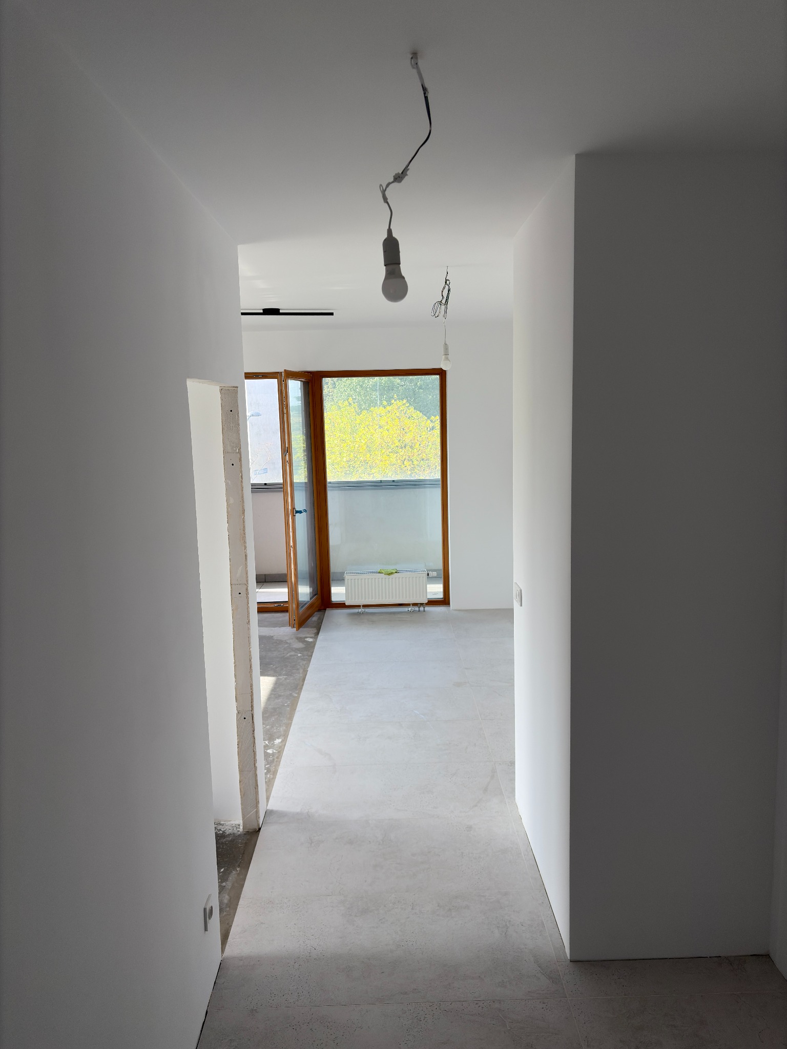 Wnętrze w trakcie wykańczania: szare płytki podłogowe, białe ściany, otwarte drewniane drzwi balkonowe z widokiem na zieleń. Wiszące żarówki bez kloszy.