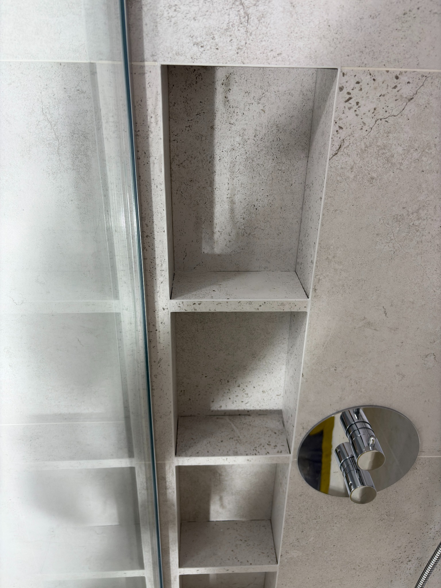 Wnęka z półkami w kabinie prysznicowej, wykończona płytkami imitującymi kamień. Widoczna szklana ściana kabiny i bateria prysznicowa.