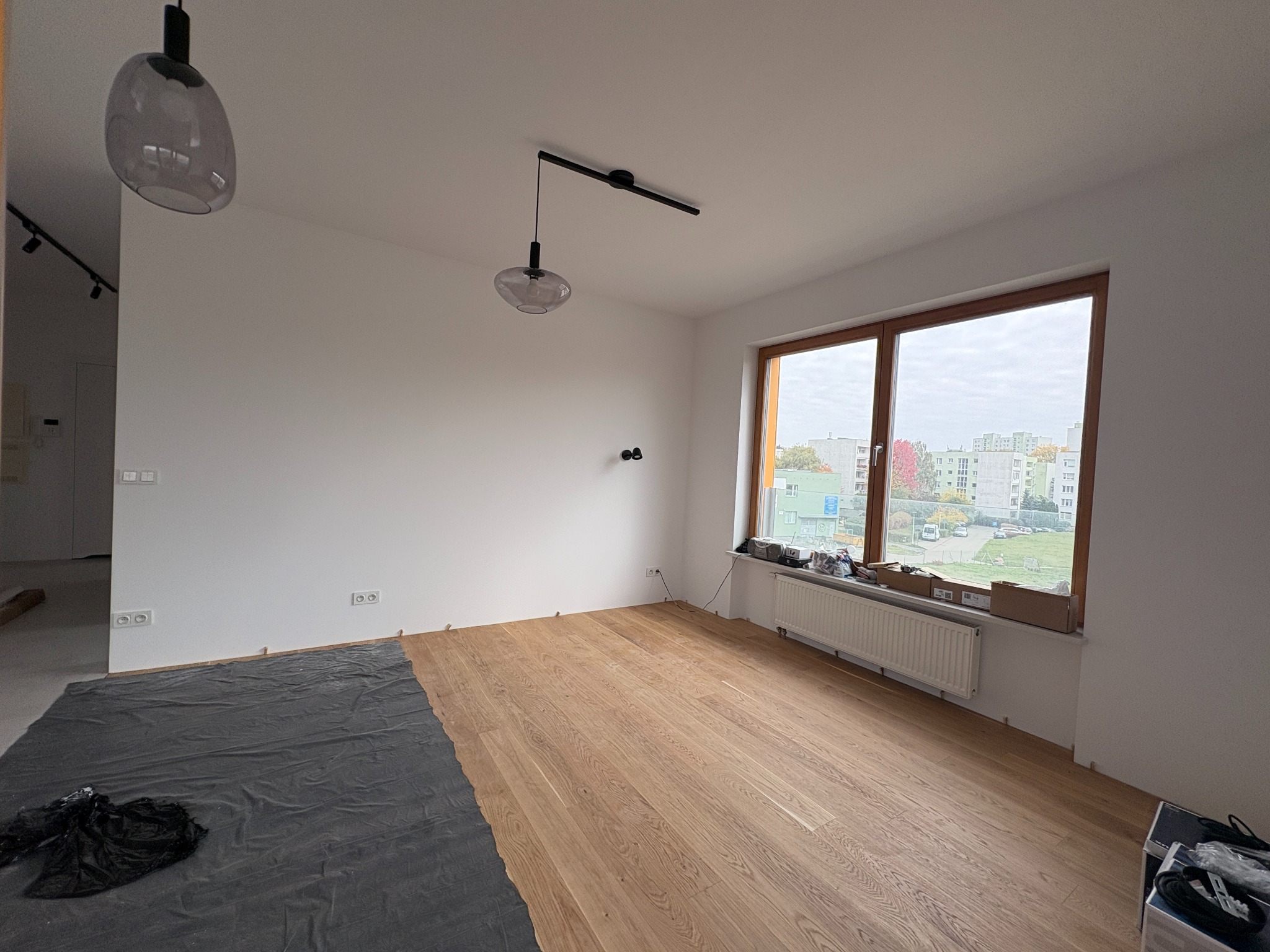 Układanie drewnianej podłogi w jasnym pokoju z dużym oknem. Część podłogi zabezpieczona folią, widoczne lampy sufitowe i grzejnik pod oknem. Prace wykończeniowe w toku.