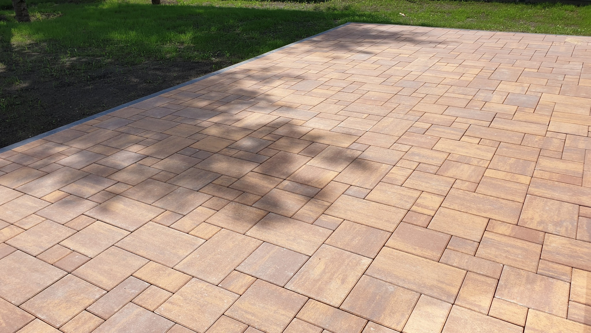 Ułożona kostka brukowa w odcieniach brązu, z nieregularnym wzorem i widocznym cieniem drzewa. W tle trawnik. Drobne nierówności w ułożeniu kostki.