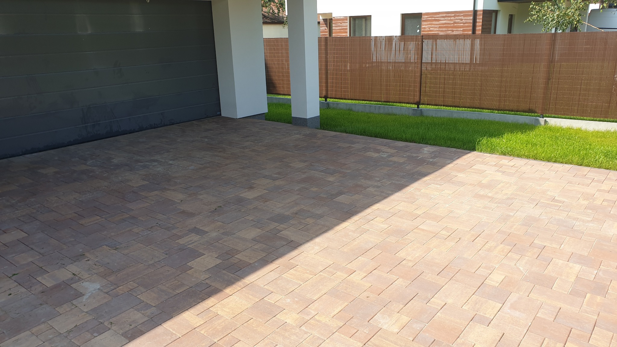 Ułożona kostka brukowa w odcieniach brązu przed garażem i ogrodzeniem z siatki, częściowo zacieniona. Widoczny fragment trawnika.