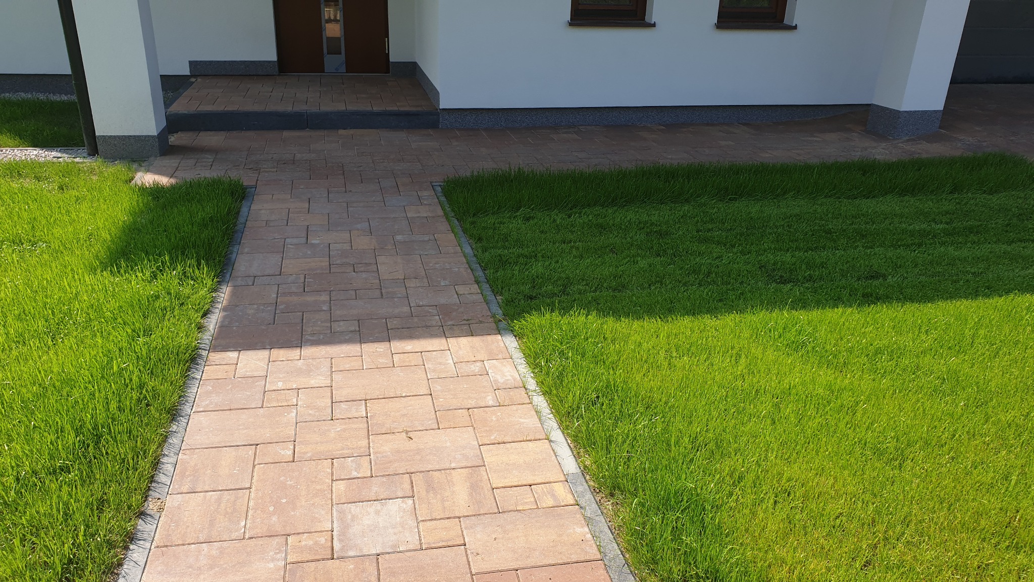 Ścieżka z kostki brukowej prowadzi do wejścia domu, otoczona świeżą, zieloną trawą. Widok na elewację i fragment drzwi wejściowych.