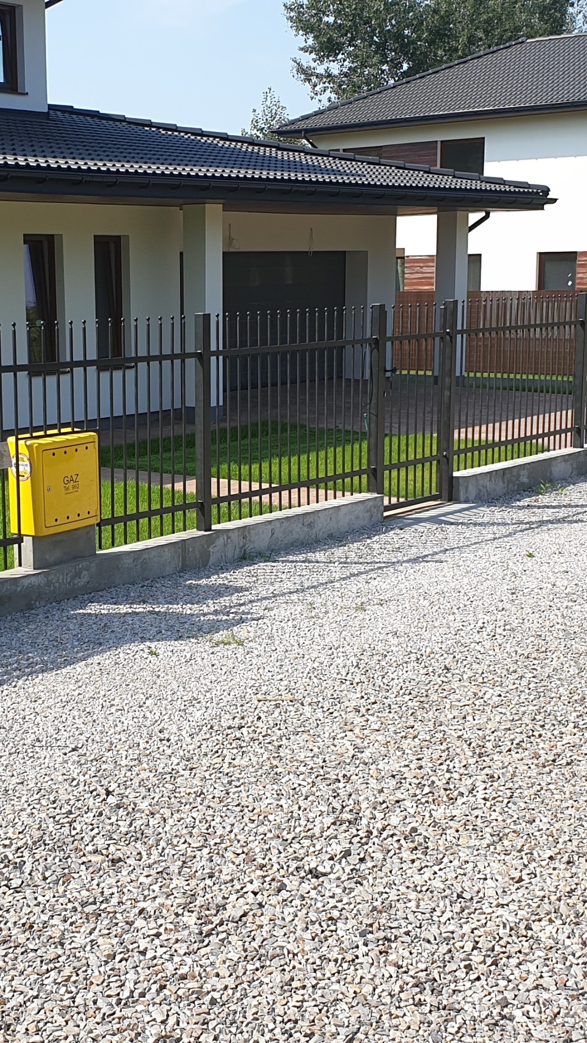 Nowoczesne ogrodzenie metalowe z bramą i furtką, betonowa podmurówka, żółta skrzynka gazowa na tle domu z garażem i podjazdem z kamienia.