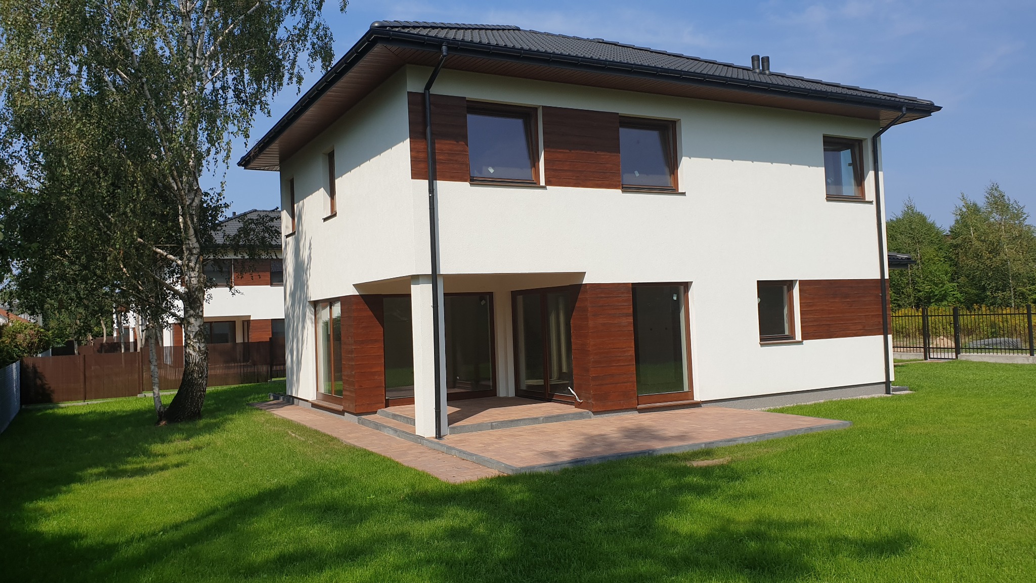 Nowoczesny dom z białymi ścianami i drewnianymi akcentami, otoczony zielonym trawnikiem i kostką brukową na tarasie. W tle widoczny fragment ogrodzenia i drzewa.