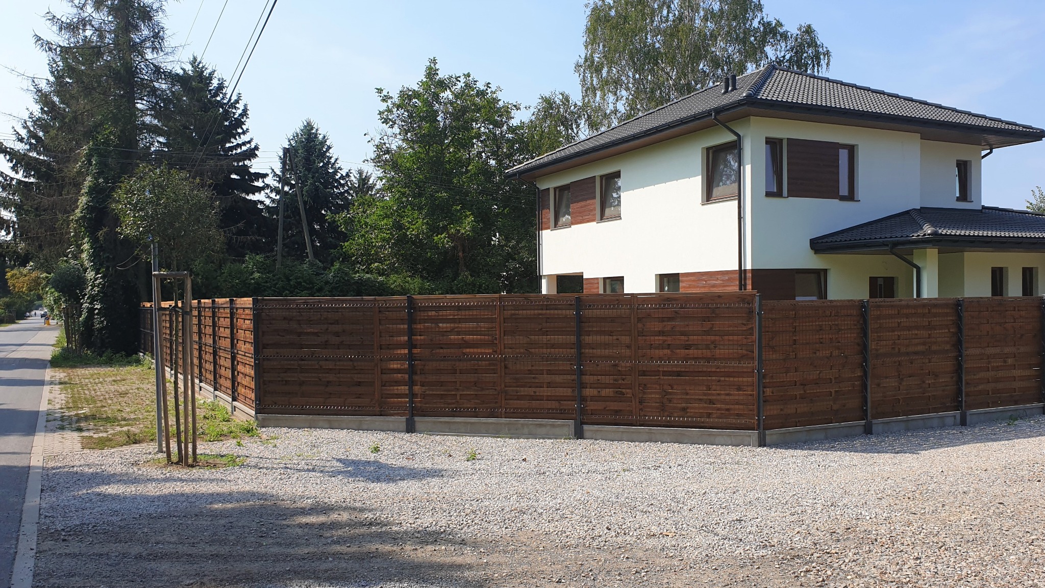 Nowoczesne ogrodzenie panelowe z poziomych desek w kolorze brązowym, na tle domu z białymi ścianami i szarym dachem, otoczone żwirowym podłożem, w słoneczny dzień.