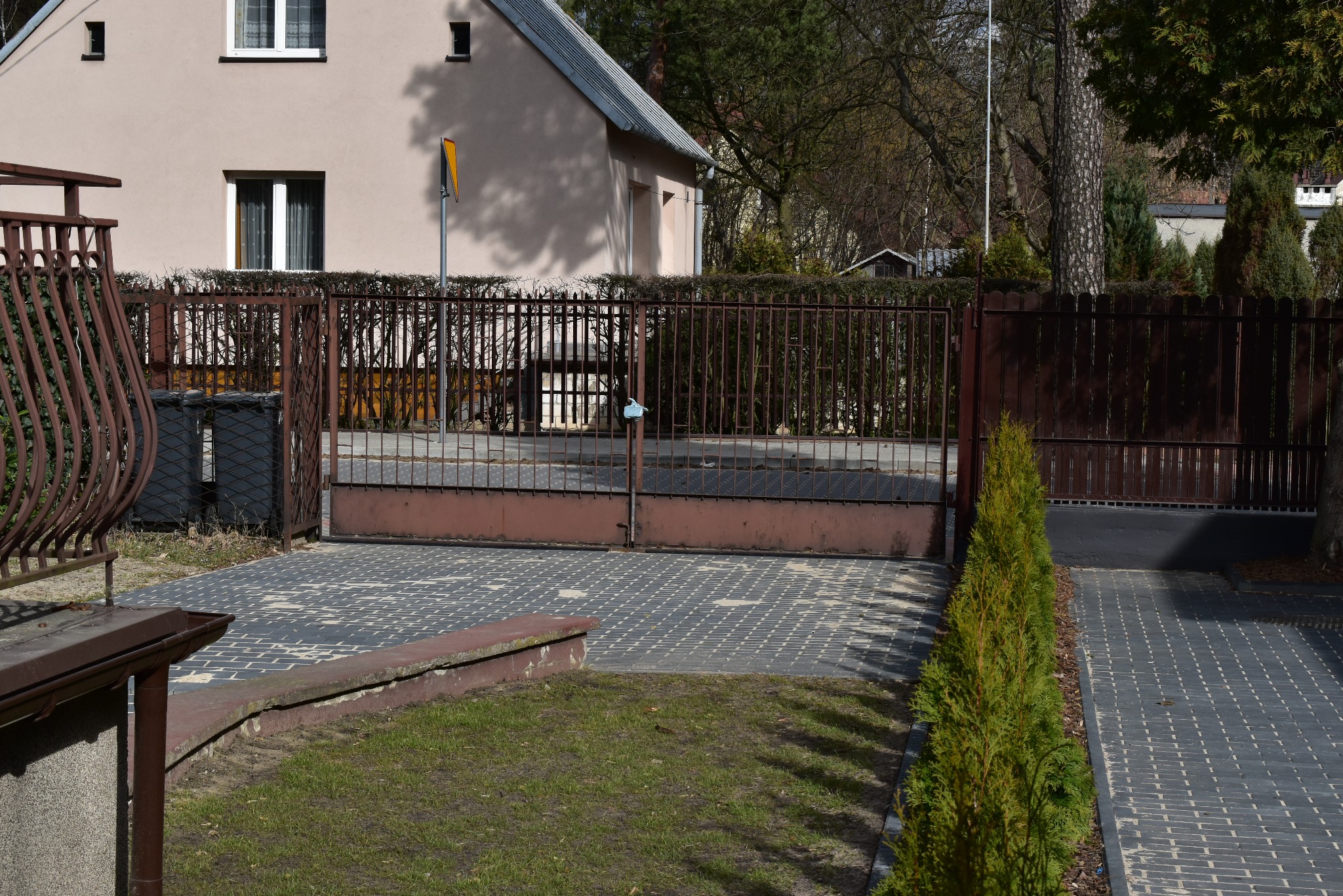 Otwarta, brązowa metalowa brama wjazdowa na posesję z kostki brukowej. W tle budynek z beżową elewacją i żywopłot. Po prawej stronie rząd małych drzewek.