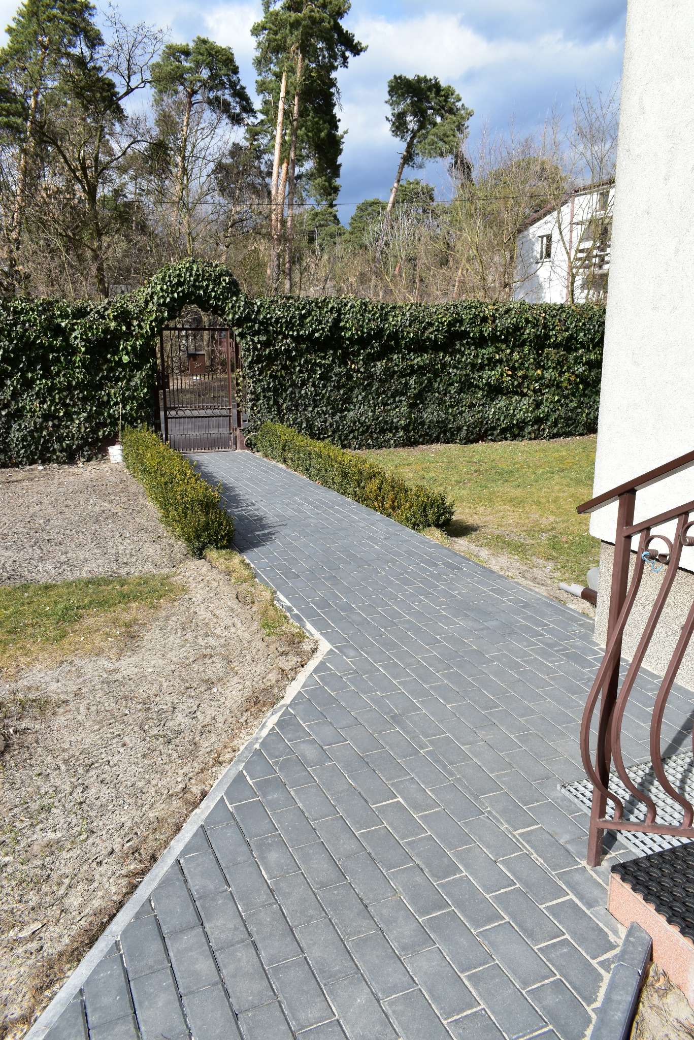 Ułożona szara kostka brukowa prowadzi do bramki w żywopłocie. Widoczne schody z metalową barierką i fragment budynku. Tło stanowią drzewa.