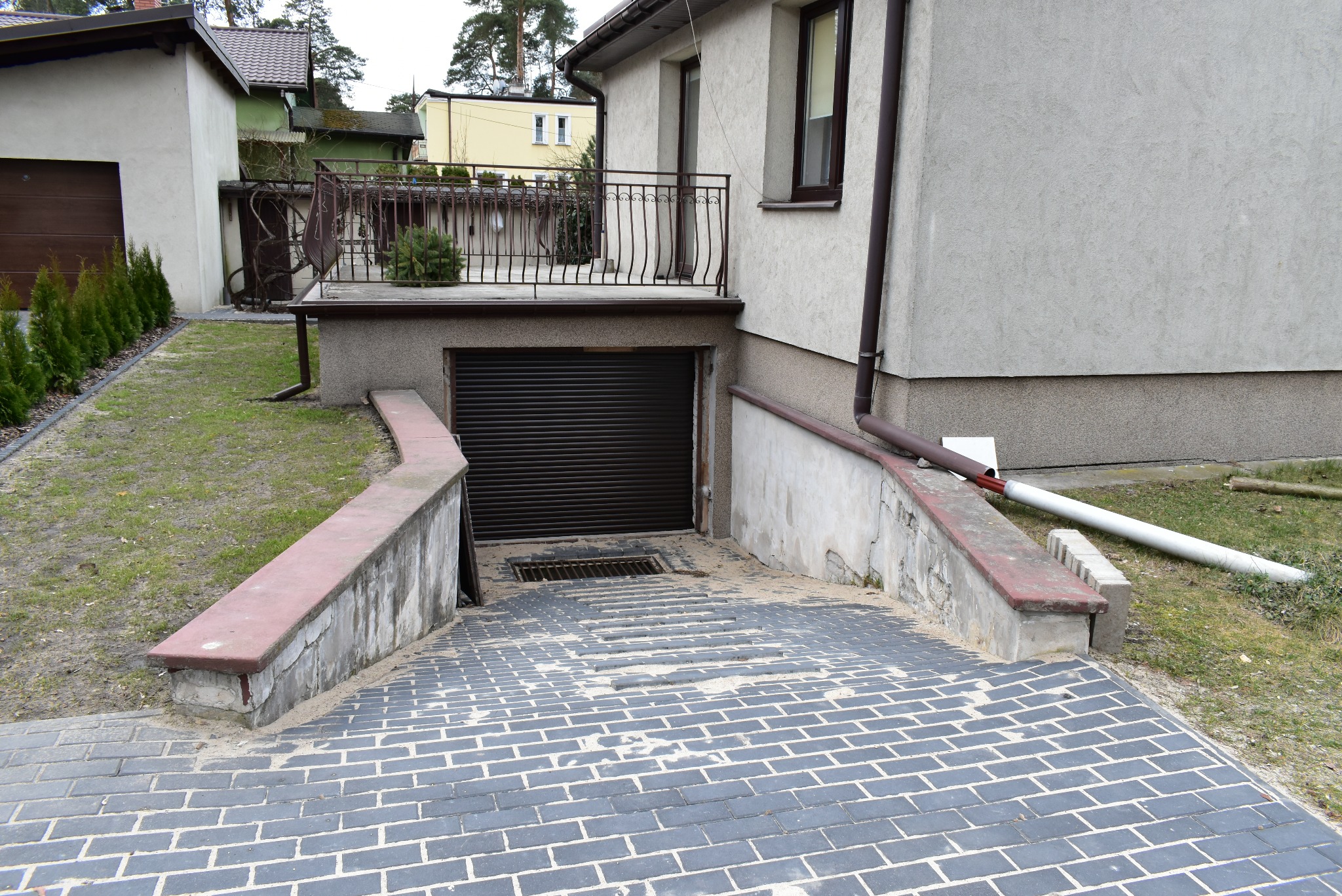 Podjazd do garażu z kostki brukowej w szarym odcieniu, z widocznym spadkiem terenu. Po bokach podjazdu znajdują się niskie murki. W tle budynek z balkonem.