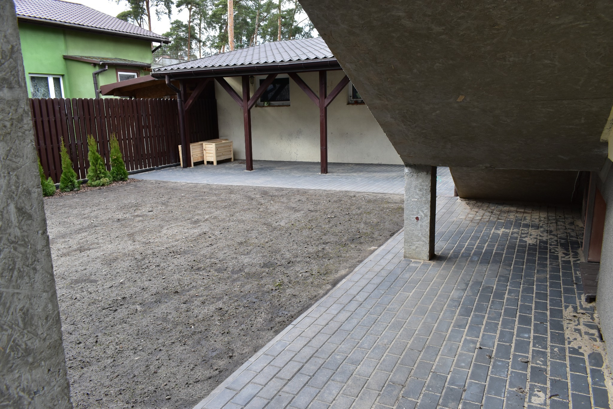 Ukończona nawierzchnia z szarej kostki brukowej przy budynku z zadaszeniem i drewnianymi słupami. W tle widoczny trawnik i ogrodzenie z zielonymi krzewami.