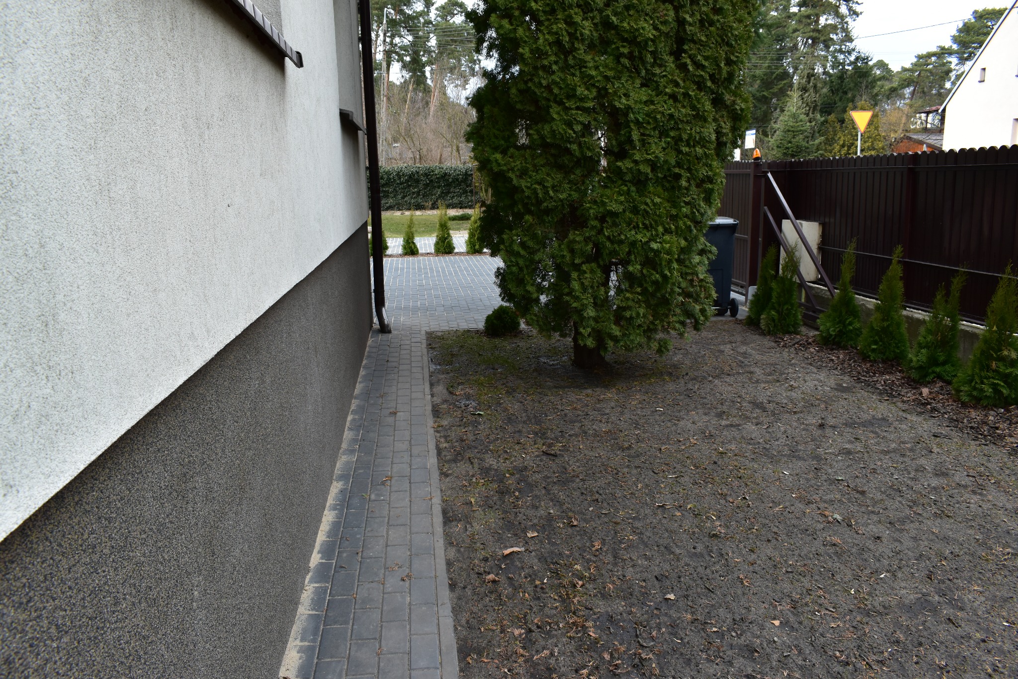 Boczny widok domu z szarą kostką brukową, ziemią i tujami. W tle brązowe ogrodzenie i drzewa. Częściowo widoczna elewacja budynku z dwoma odcieniami szarości.