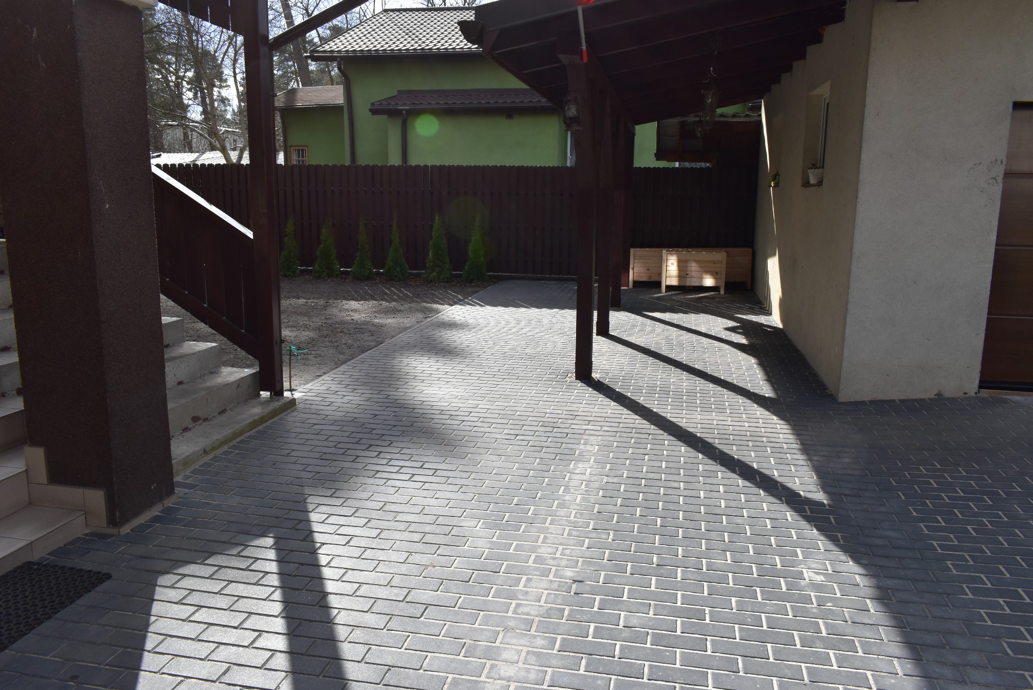 Szara kostka brukowa w regularnym wzorze, oświetlona słońcem, z widocznymi elementami architektury domu i ogrodu w tle. Widok na podjazd.