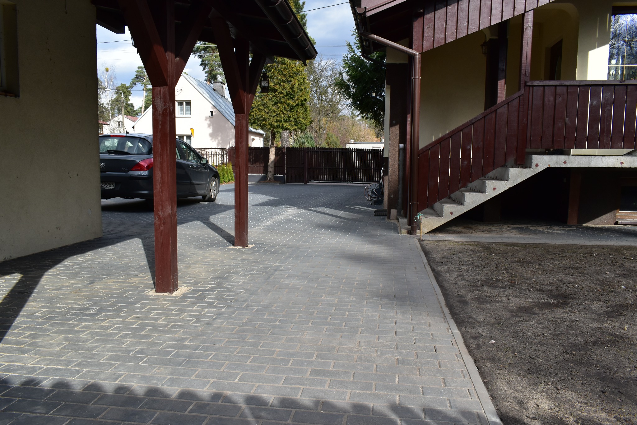 Szara kostka brukowa ułożona na podjeździe z zadaszeniem i schodami. Widoczny samochód i ogrodzenie w tle. Ciemne filary zadaszenia rzucają cień na kostkę.