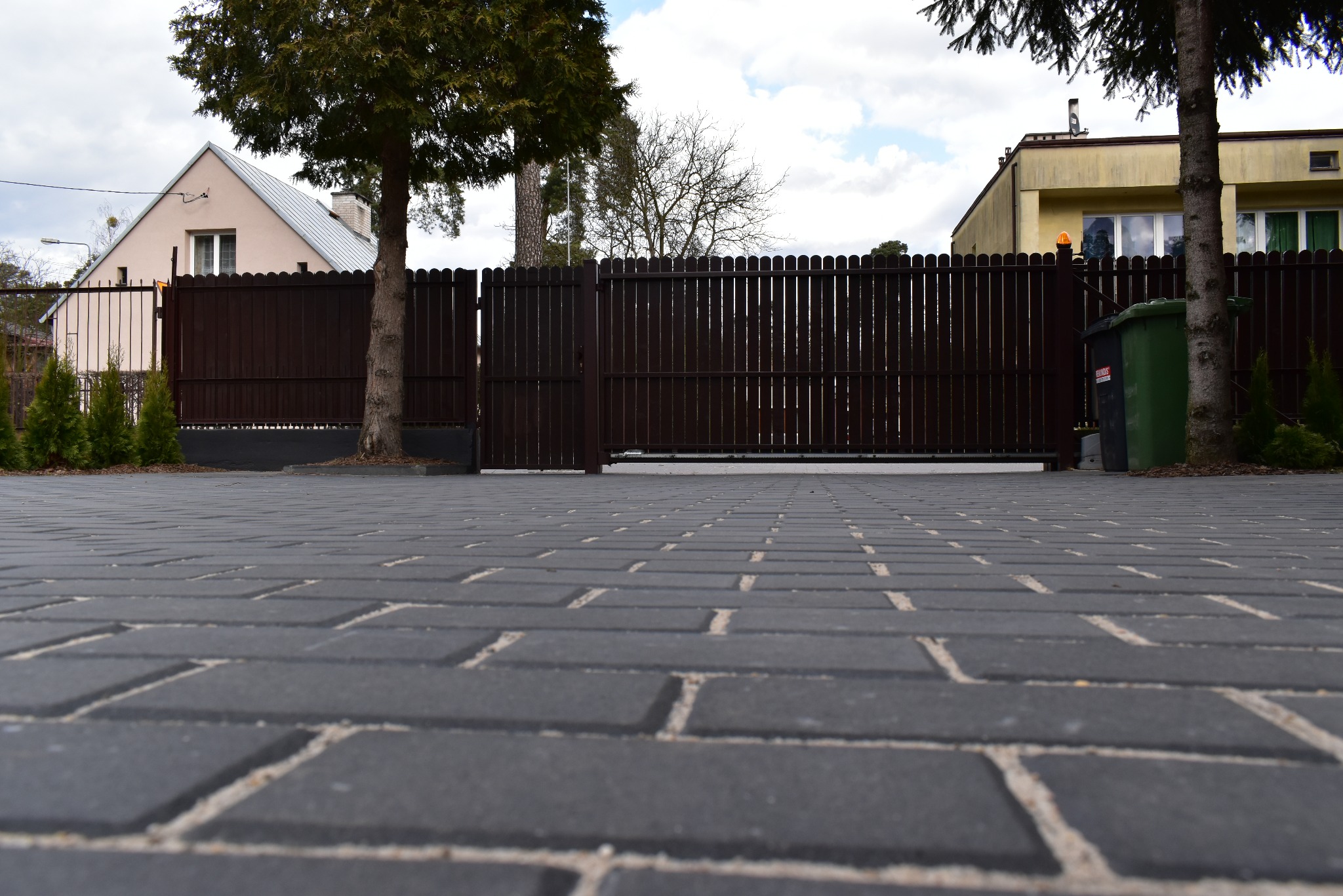 Ciemnoszara kostka brukowa prowadząca do brązowej, pionowej bramy wjazdowej z drewnianymi panelami, otoczonej drzewami i domami w tle.