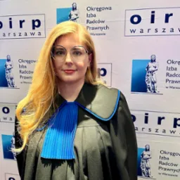 Portret kobiety w todze radcowskiej na tle baneru Okręgowej Izby Radców Prawnych w Warszawie. Uśmiechnięta, w okularach, z rozpuszczonymi blond włosami.
