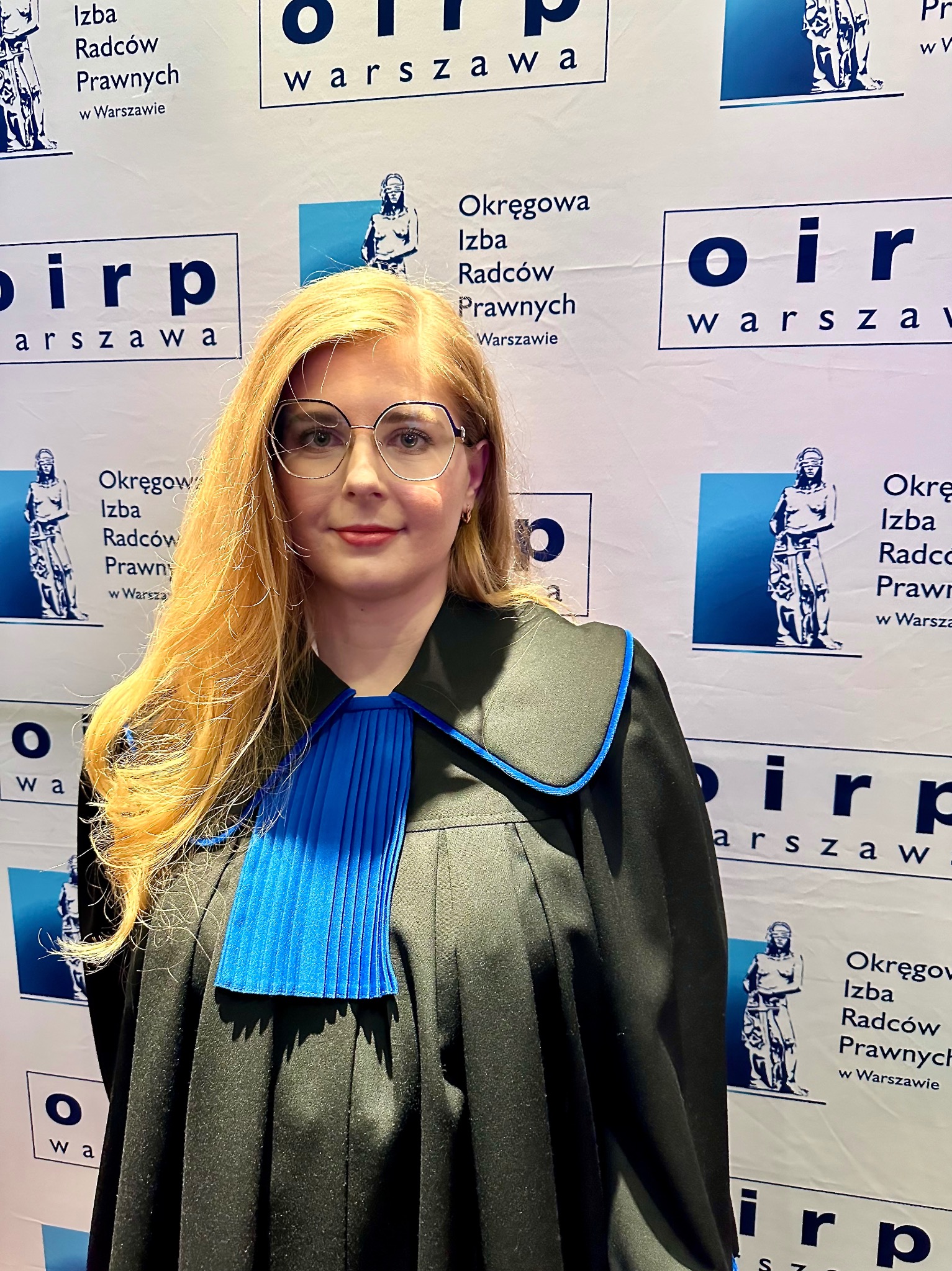 Portret kobiety w todze radcowskiej na tle baneru Okręgowej Izby Radców Prawnych w Warszawie. Uśmiechnięta, w okularach, z rozpuszczonymi blond włosami.