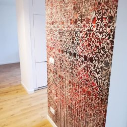 OLEG PANKIV USŁUGI WYKOŃCZENIOWE - Wnętrze po wykończeniu: wzorzysta tapeta w odcieniach czerwieni i brązu na ścianie, jasna podłoga imitująca drewno, białe listwy przypodłogowe i fragment szafy wnękowej.