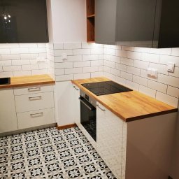 OLEG PANKIV USŁUGI WYKOŃCZENIOWE - Nowoczesna kuchnia po remoncie: białe szafki z drewnianym blatem, czarna płyta indukcyjna i piekarnik. Wzorzysta, czarno-biała podłoga i białe kafelki na ścianie.