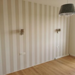 OLEG PANKIV USŁUGI WYKOŃCZENIOWE - Pokój po malowaniu ścian w pionowe pasy beżu i bieli, z drewnianą podłogą, kinkietami i lampą pod sufitem owiniętą folią ochronną.