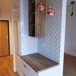 OLEG PANKIV USŁUGI WYKOŃCZENIOWE - Biała zabudowa meblowa z drewnianym blatem i półkami, akcentowana miedzianymi lampami wiszącymi na tle ściany w biało-szarym wzorze jodełki.