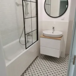 Nowoczesna łazienka z czarno-białą mozaiką podłogową, kabiną prysznicową w stylu loft i okrągłym lustrem nad umywalką na szafce z drewnianym blatem.