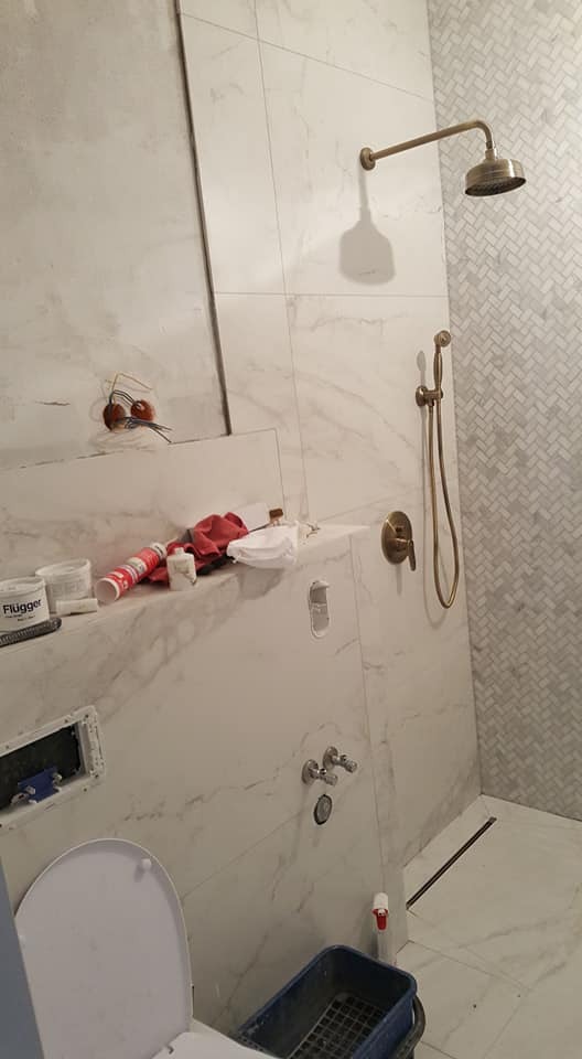 Wnętrze łazienki w trakcie wykańczania: białe płytki imitujące marmur, złota armatura prysznicowa, widoczne narzędzia i materiały budowlane, mozaika w jodełkę.