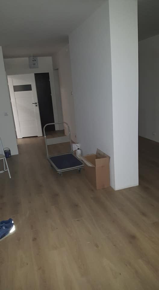 Jasne panele podłogowe w nowym mieszkaniu. W tle biała ściana, drzwi i wózek transportowy. Widok na otwarte pomieszczenia.