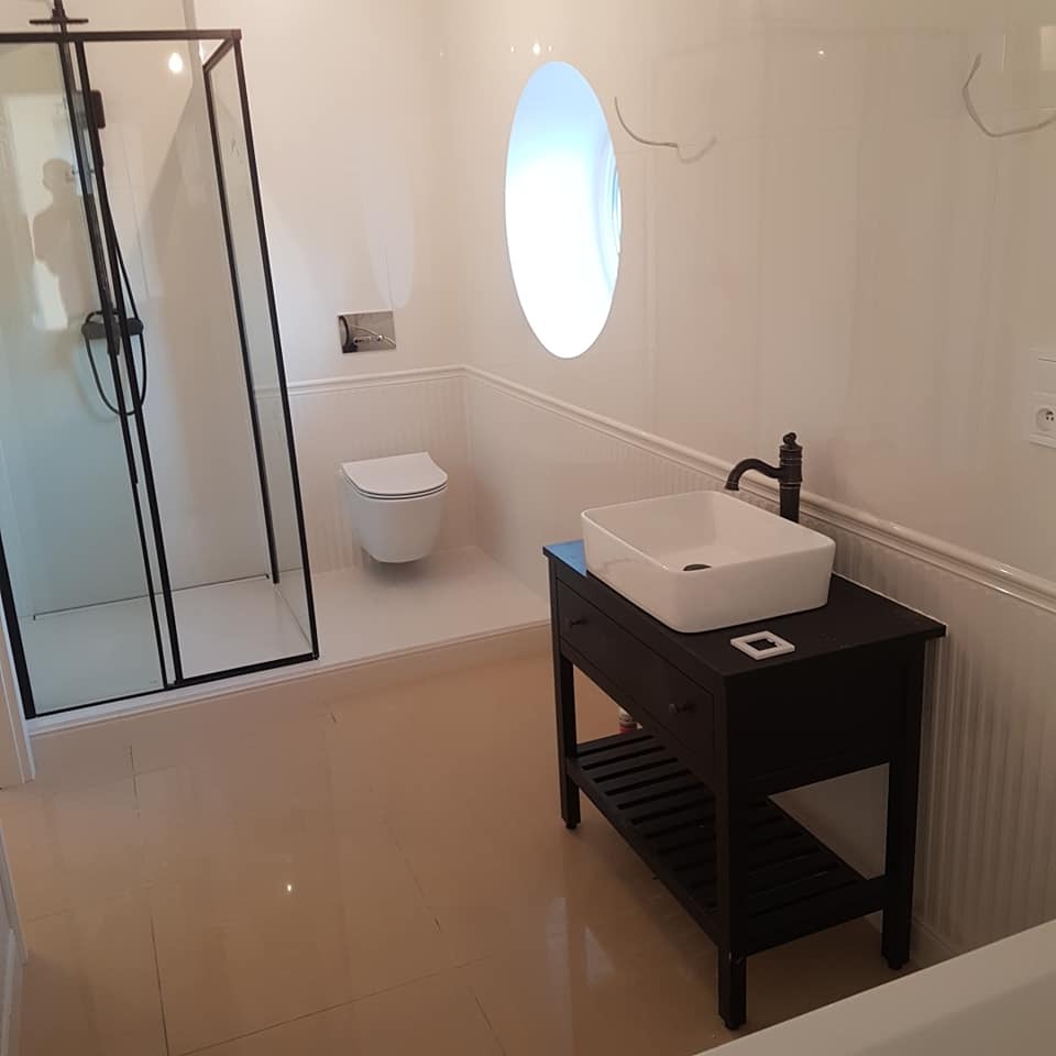 Biała łazienka z czarną kabiną prysznicową i umywalką na szafce. Okrągłe okno nadaje wnętrzu unikalny charakter.