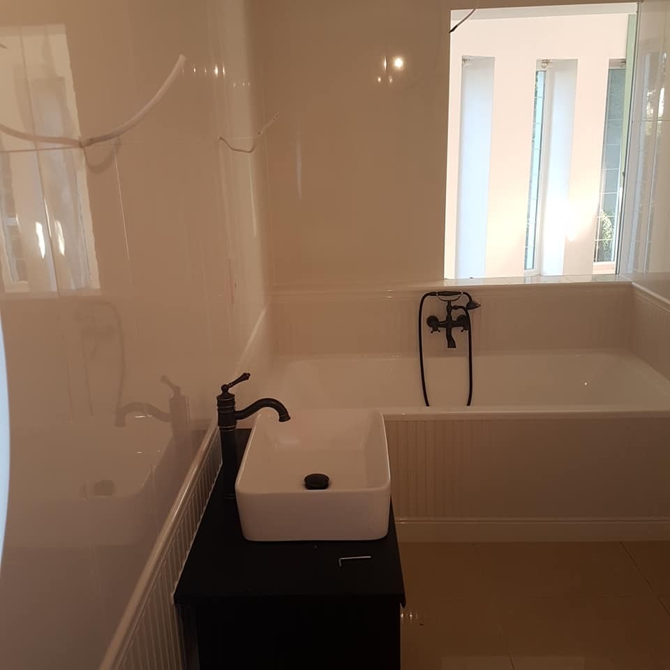 Biała łazienka z wanną i umywalką w stylu retro z czarną armaturą. Ściany wykończone panelami, okno z widokiem na zieleń.