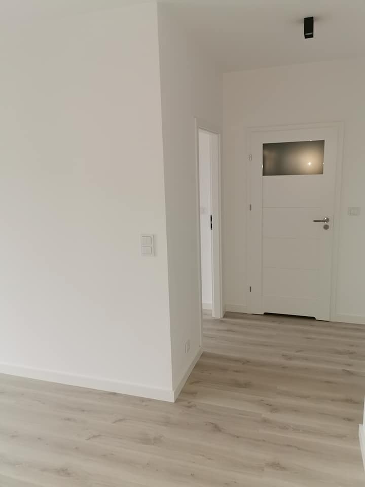 Jasne wnętrze po remoncie: białe ściany, drewnopodobna podłoga, minimalistyczne drzwi z przeszkleniem i czarny punktowy reflektor sufitowy.