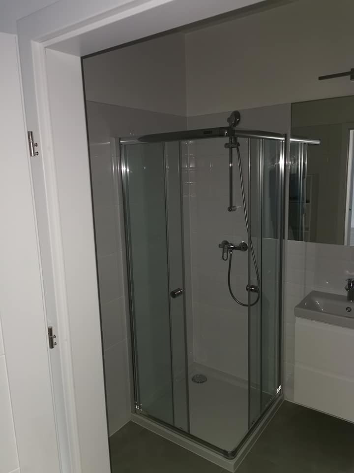 Nowoczesna kabina prysznicowa z chromowanymi detalami w wyremontowanej łazience. Widoczne płytki i umywalka. Minimalistyczny design i jasne kolory.