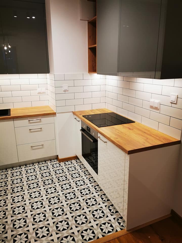 Nowoczesna kuchnia po remoncie: białe szafki z drewnianym blatem, czarna płyta indukcyjna i piekarnik. Wzorzysta, czarno-biała podłoga i białe kafelki na ścianie.