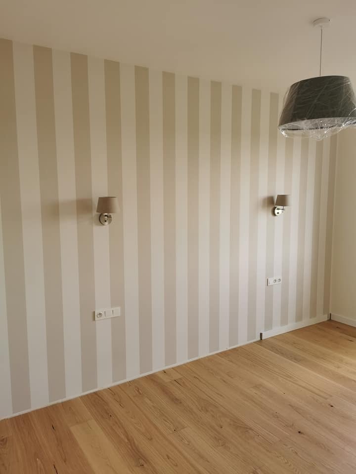 Pokój po malowaniu ścian w pionowe pasy beżu i bieli, z drewnianą podłogą, kinkietami i lampą pod sufitem owiniętą folią ochronną.
