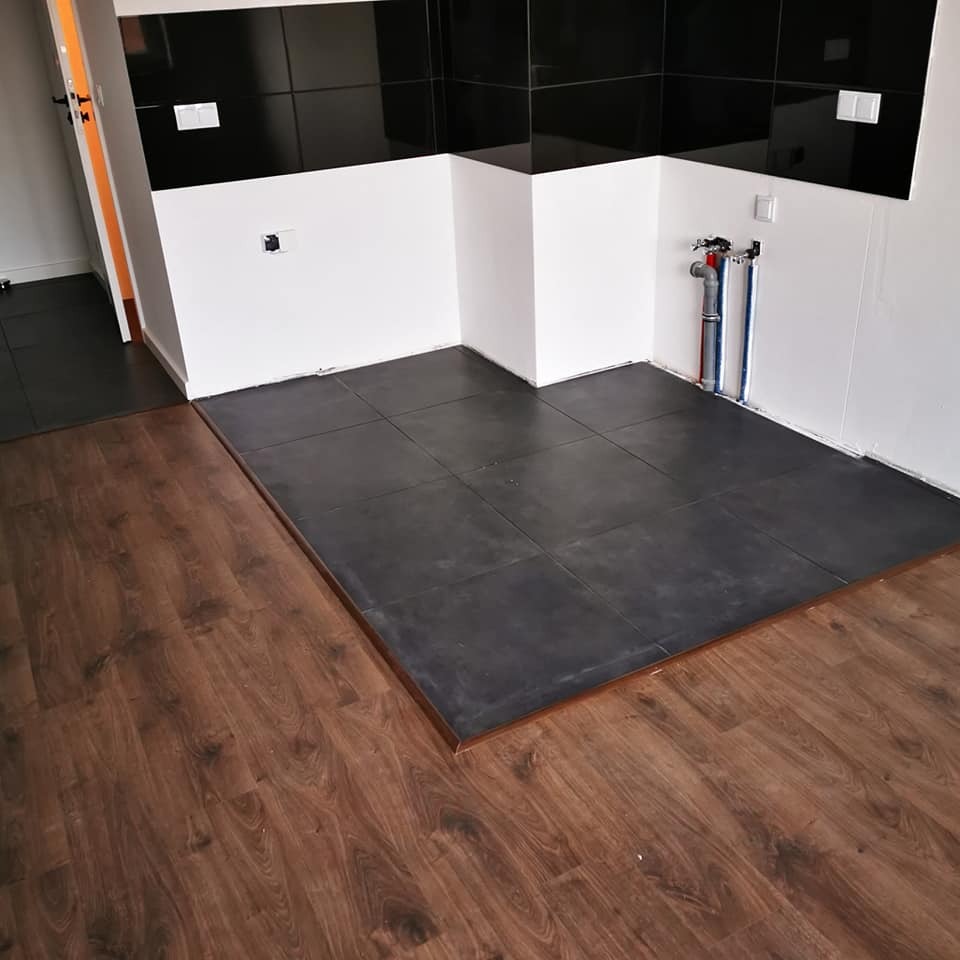 Ciemne płytki podłogowe w łazience, ułożone na podwyższeniu z drewnopodobną listwą, ściany w bieli i czerni. Widoczne rury instalacji wodnej.