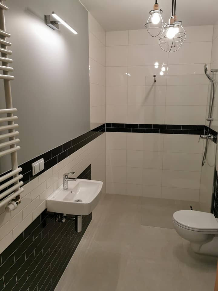 Nowoczesna łazienka z biało-czarnymi płytkami, wiszącą umywalką i grzejnikiem drabinkowym. Minimalistyczne lampy sufitowe i szara ściana dodają elegancji.