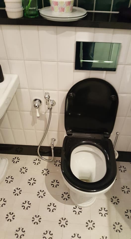 Toaleta z czarną deską i bidetką na tle białych płytek z wzorem. Nad toaletą lustro i półka z dekoracjami. Ujęcie z góry, prezentujące wykończoną łazienkę.