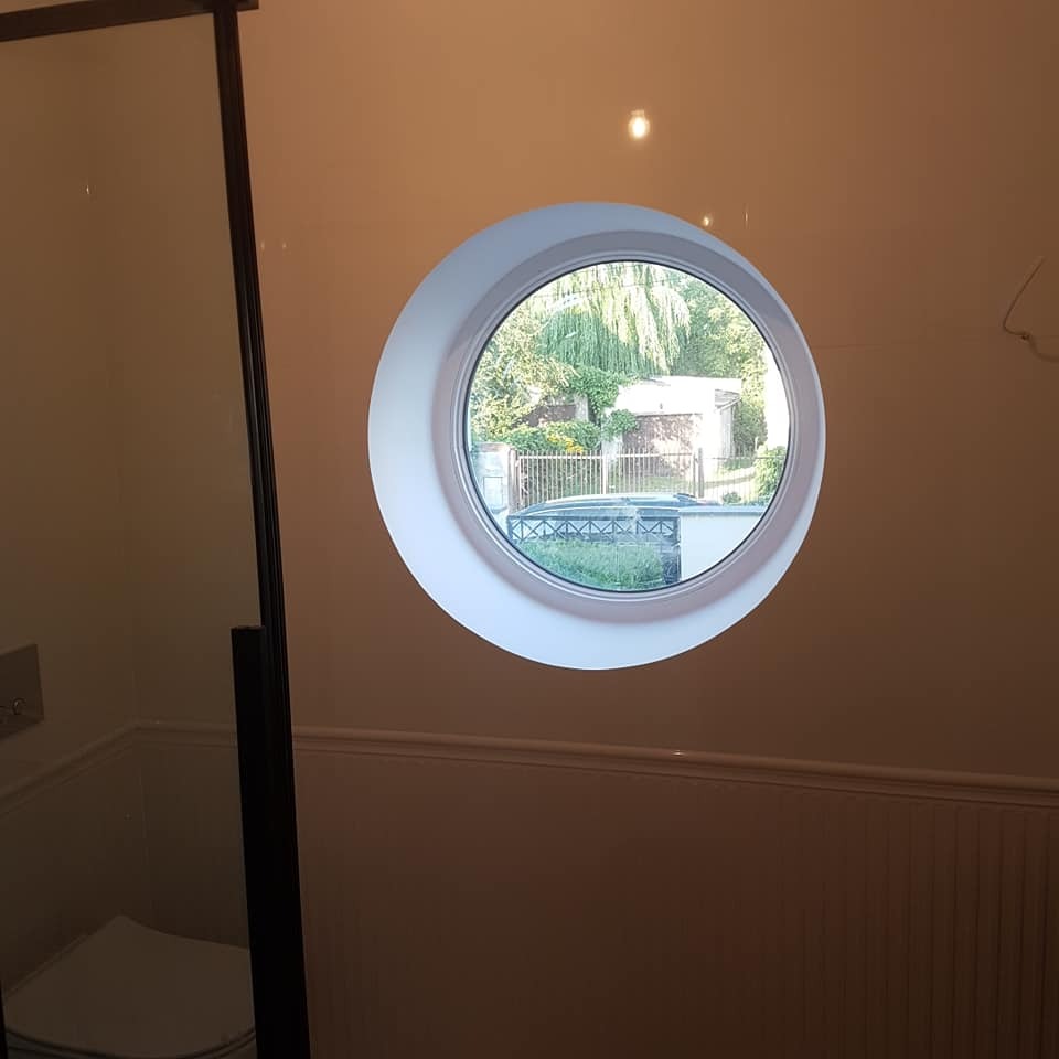 Jasna łazienka z okrągłym oknem z widokiem na ogród. Kabina prysznicowa z czarną ramą po lewej stronie. Ściany w odcieniach beżu z dekoracyjną listwą.