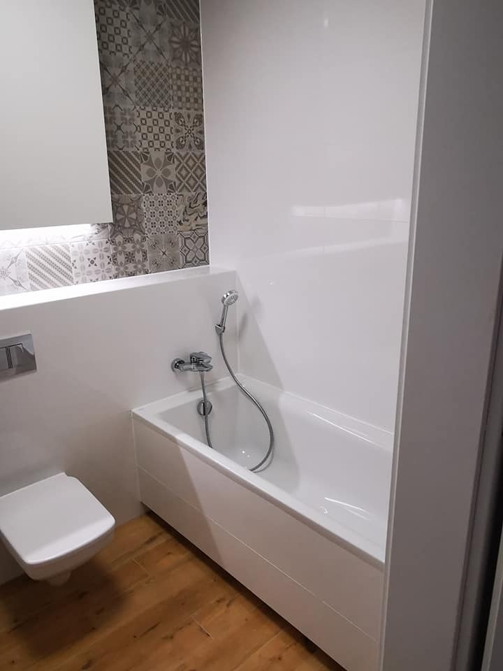 Biała wanna z prysznicem i toaleta w wykończonej łazience. Na ścianie dekoracyjne płytki z wzorem. Podłoga z paneli imitujących drewno.