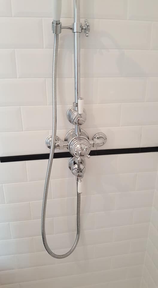 Bateria prysznicowa chromowana z białą rączką na tle białych płytek ułożonych w cegiełkę, z czarnym akcentem dekoracyjnym. Detal wykończenia łazienki.