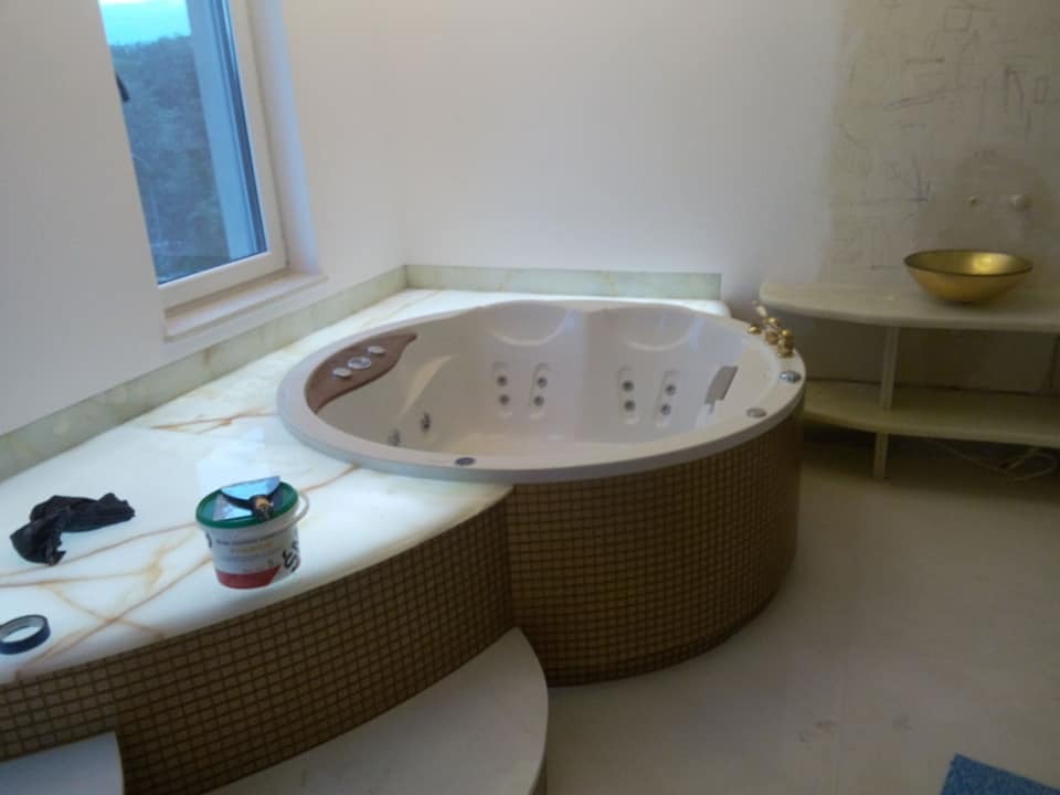 Owalna wanna z hydromasażem, obudowana mozaiką, osadzona w blacie z kamienia. Widoczne akcesoria montażowe i umywalka na półce. Jasne wnętrze łazienki.