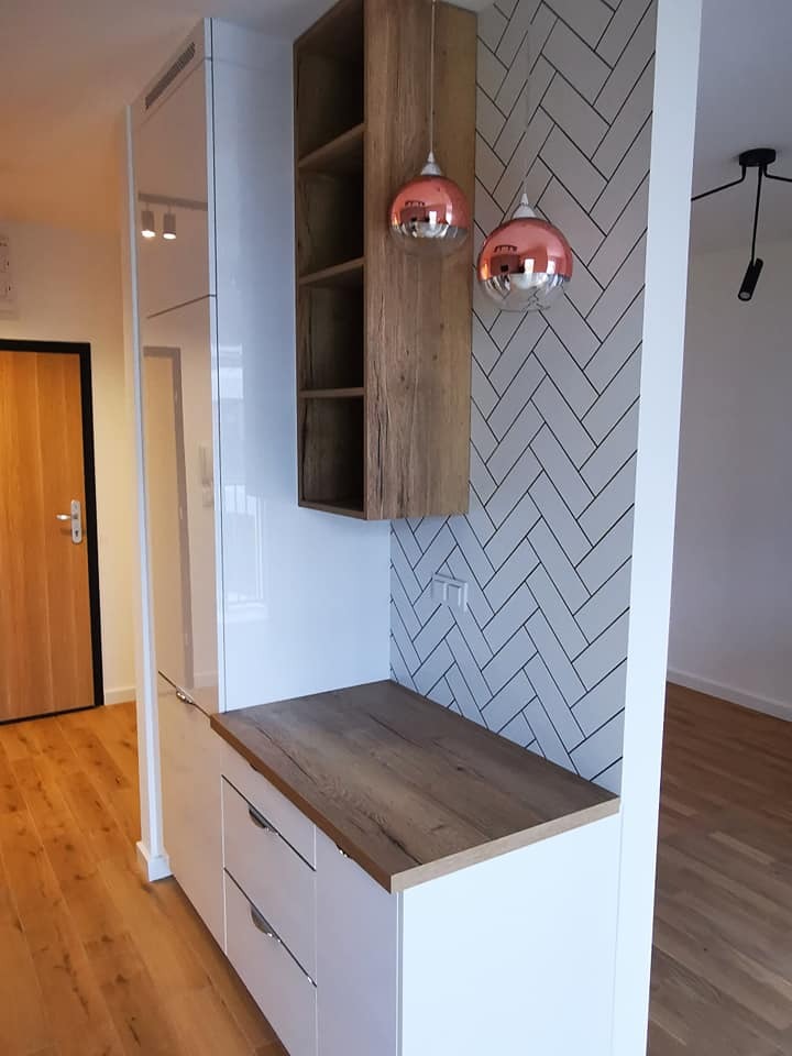 Biała zabudowa meblowa z drewnianym blatem i półkami, akcentowana miedzianymi lampami wiszącymi na tle ściany w biało-szarym wzorze jodełki.