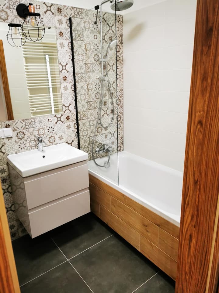 Nowoczesna łazienka z wiszącą szafką, mozaiką nad wanną i szklanym parawanem. Ciemna podłoga i designerska lampa dodają wnętrzu charakteru.