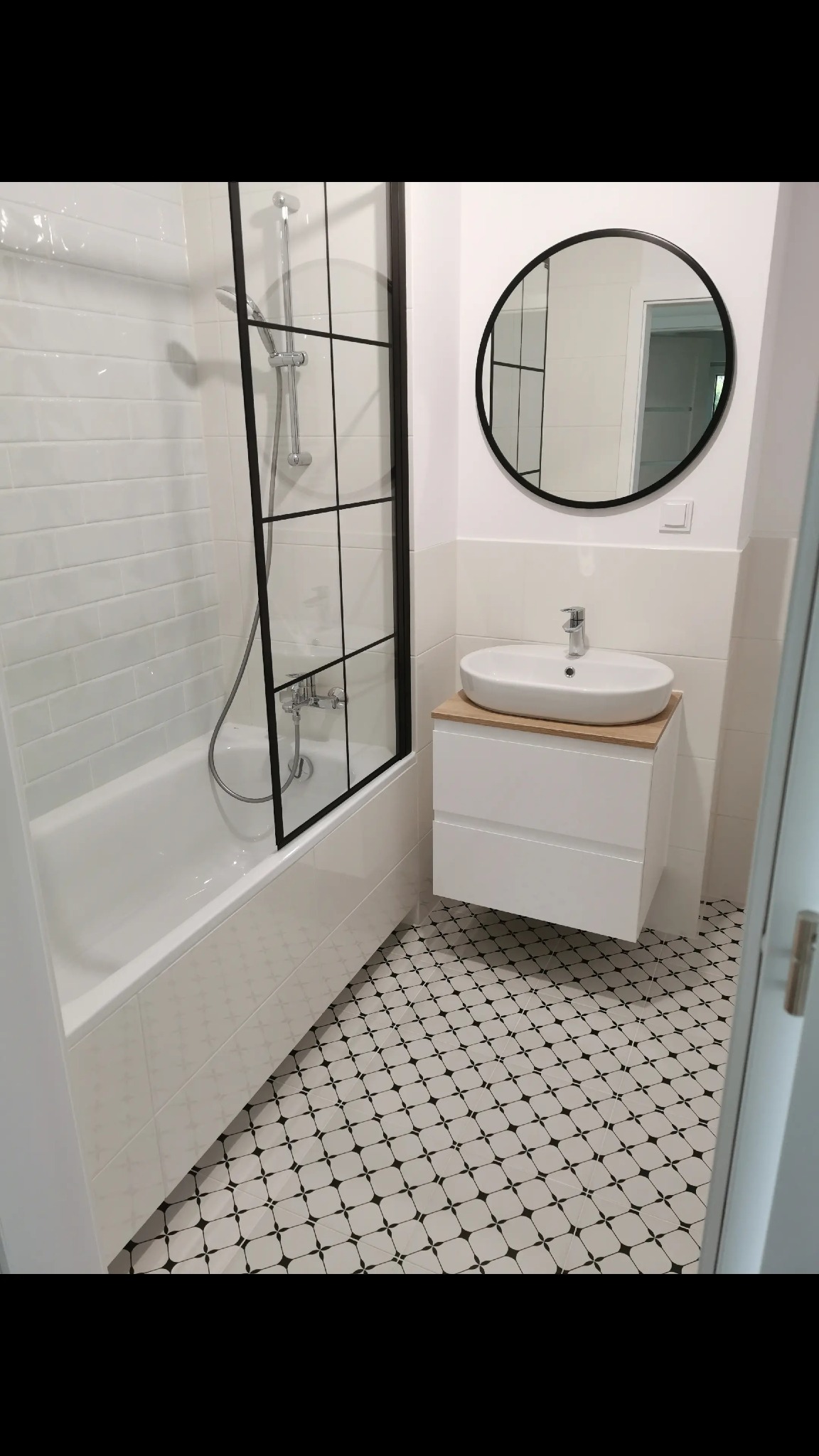 Nowoczesna łazienka z czarno-białą mozaiką podłogową, kabiną prysznicową w stylu loft i okrągłym lustrem nad umywalką na szafce z drewnianym blatem.