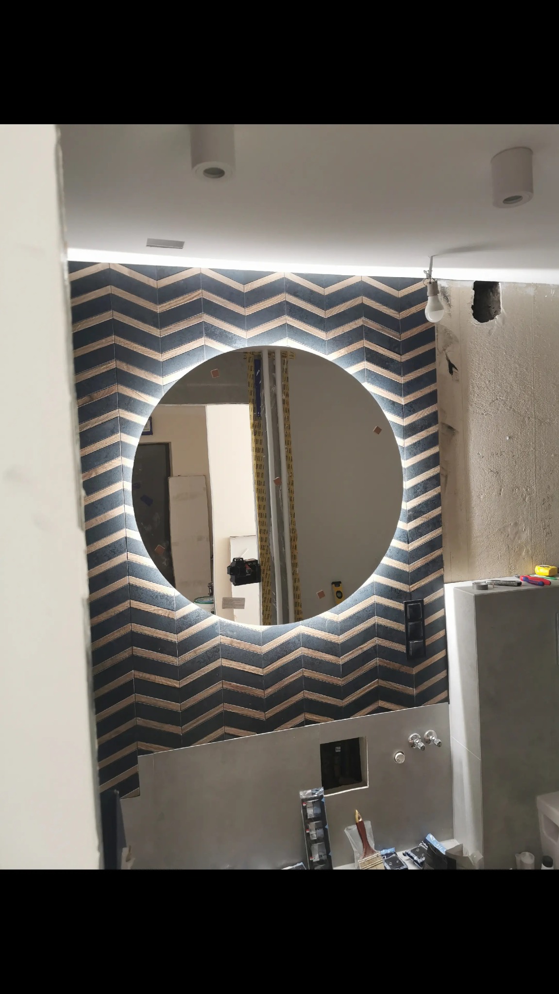 Łazienka w trakcie remontu: okrągłe lustro LED na ścianie z chevronowym wzorem płytek, widoczne narzędzia i niedokończone elementy instalacji.