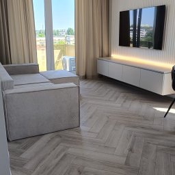 Oddział Czysta Sprawa - Jasny salon z parkietem w jodełkę, beżową sofą i ścianą z telewizorem podświetlanym ledami. Widok na miasto przez okno balkonowe.