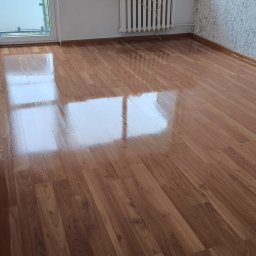 Oddział Czysta Sprawa - Błyszcząca, świeżo umyta podłoga z paneli w pokoju z oknem i grzejnikiem. Widoczne odbicia w podłodze. Tapeta w delikatny wzór na ścianie.