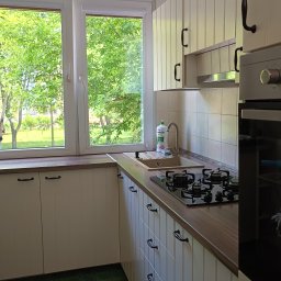 Oddział Czysta Sprawa - Jasna kuchnia z oknem na zieleń. Białe szafki z czarnymi uchwytami, zielona podłoga. Na blacie kuchennym zlew i płyta gazowa. Widok na drzewa za oknem.